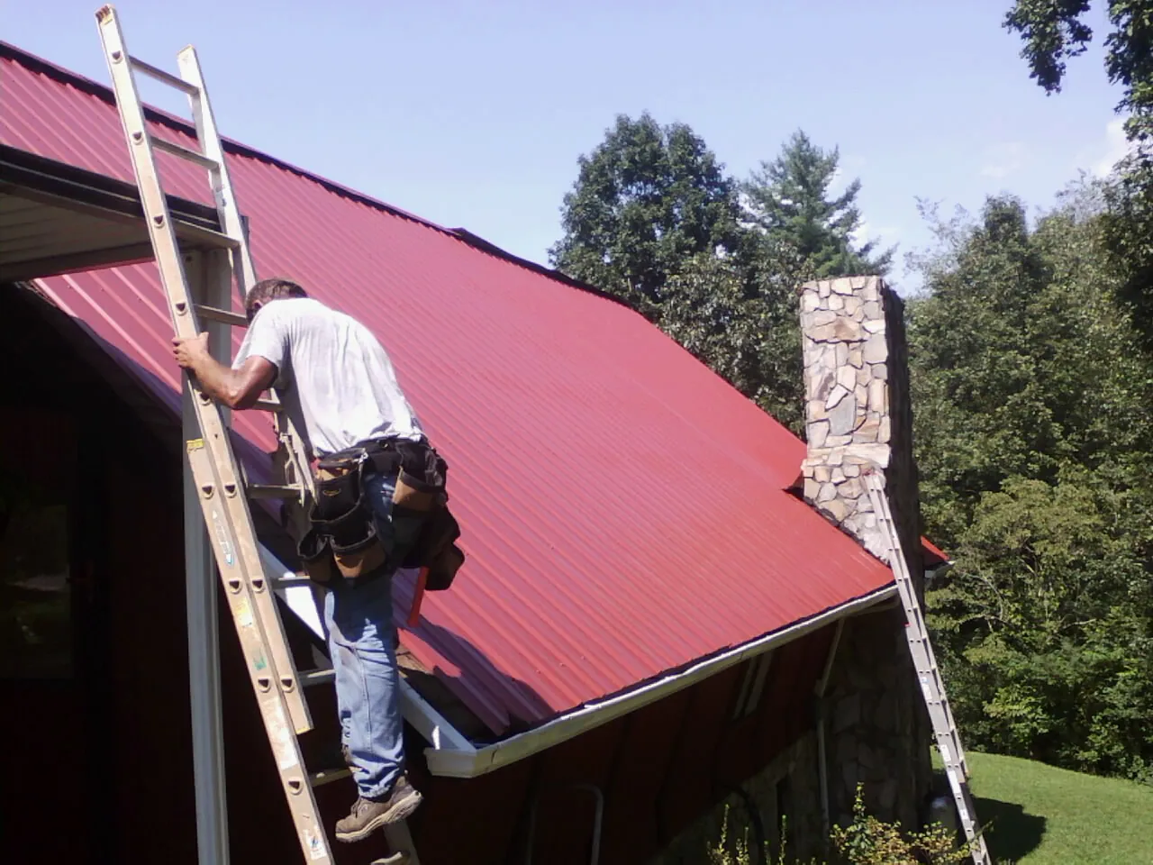 Roofer on a ladder beside a red metal roof and stone chimney — Soffit & Fascia Repair by Roofing Brothers in Melvin